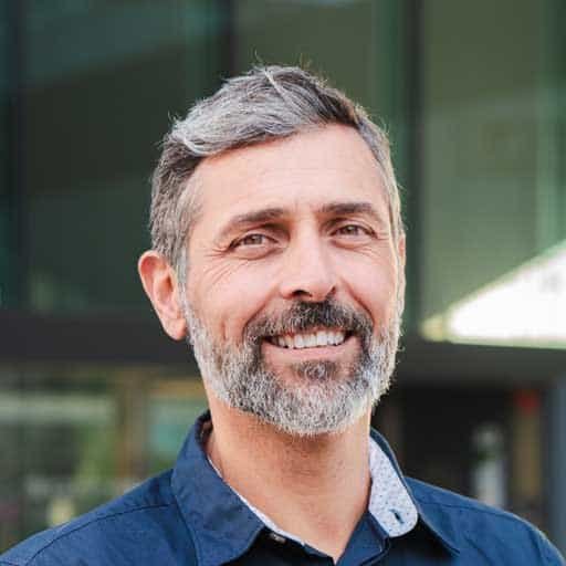 A smiling middle-aged man with gray hair and beard, wearing a blue shirt, standing outdoors in front.